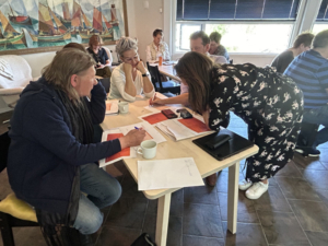 En kvinne står bøyd over et bord og skriver på et ark. Resten av rommet er fylt med personer som sitter å jobber. Det er ark og kaffekopper på bordene. I bakgrunnen er et vindu som er halveis åpent. 